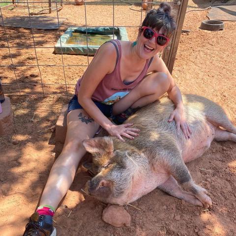 A woman is smiling, wearing pink sunglasses and sitting on the dirt ground while petting a sleeping pig.